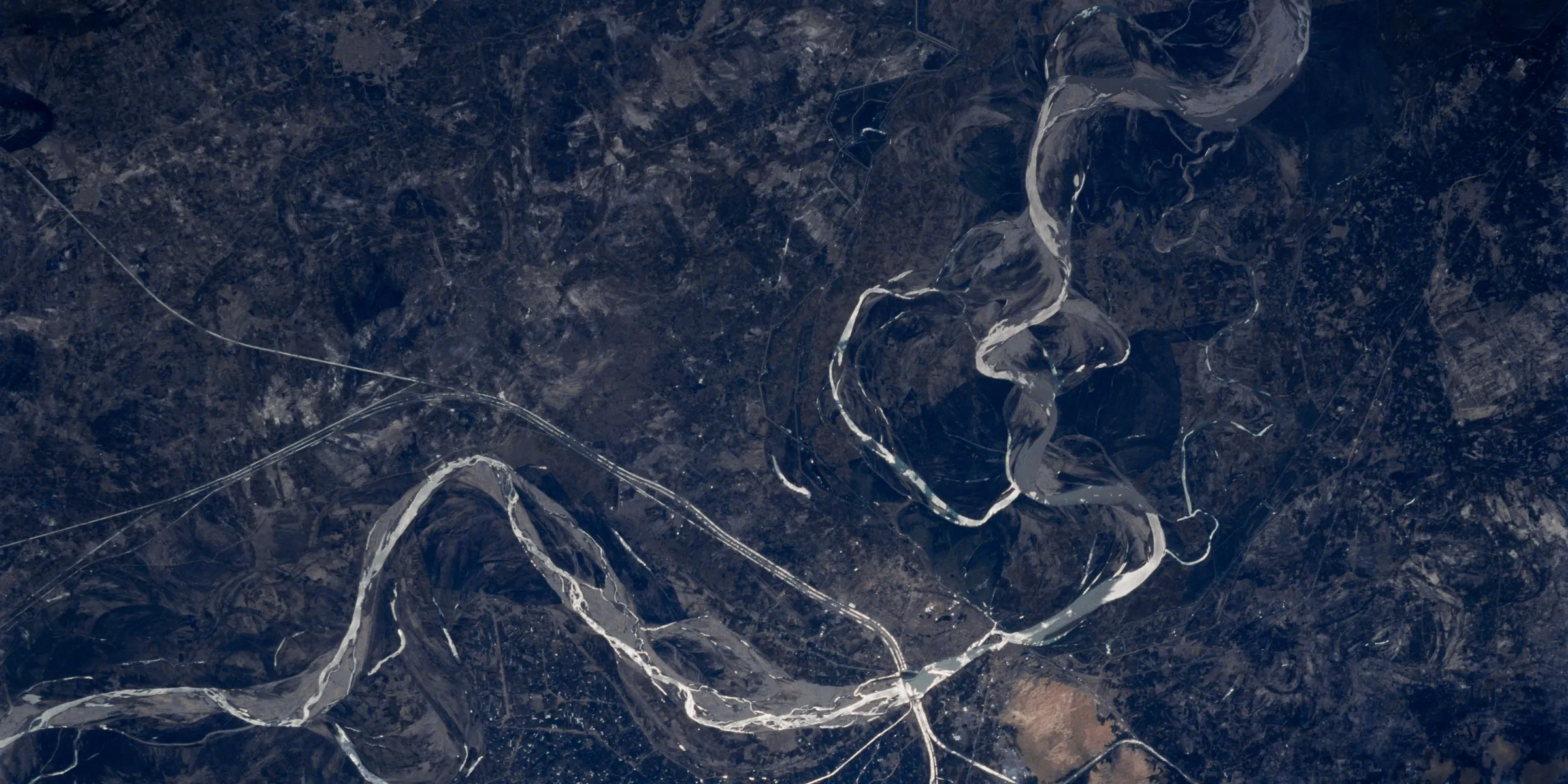 The Indus River photographed from orbit in sunglint: a bright meander crossing a dark alluvial plain, cut along a seam with its right side raised.